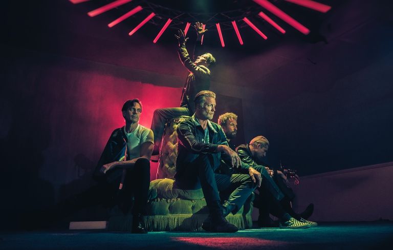 Rock band Queens of the Stone Age pose in dramatic red light, featuring frontman Josh Homme sitting front and center with bandmates.